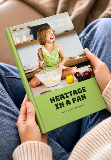 Hands holding a green Heritage in a Pan cookbook in a lap